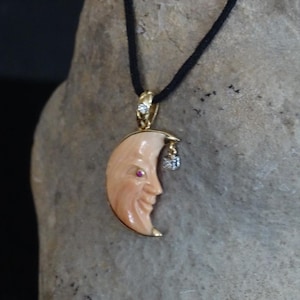 May include: A gold pendant necklace featuring a carved moon with a face, a small red gem, and a dangling charm. The moon pendant is suspended from a black cord. The pendant is displayed against a textured, neutral background.
