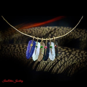 May include: A gold necklace with a row of seven gemstone feather pendants. The feathers are carved from different colored stones, including blue, white, brown, green, and black. Each feather has a small gemstone set in the center.