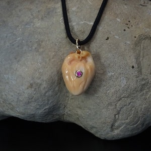May include: A carved heart pendant necklace with a pink gemstone in the center. The pendant is made of a light brown material and is hanging from a black cord.