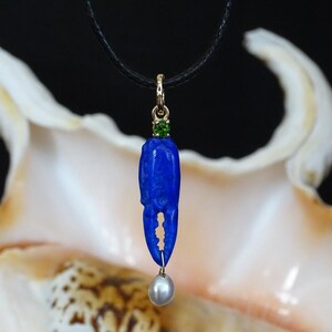 Op de afbeelding: Een blauwe lapis lazuli krabbenklauw hanger met groene edelsteen accenten en een parel die eronder hangt. De hanger zit aan een zwart koord.