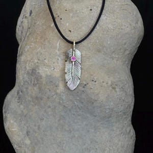 May include: A silver feather pendant necklace featuring a small, round, pink gemstone set in the center. The pendant hangs from a thin, black cord, showcasing detailed etched lines along the surface of the metallic feather design.