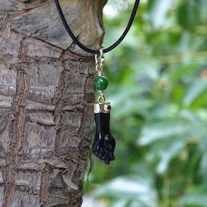 Op de afbeelding: Zwarte hangerketting met een gedetailleerde kleine hand. De hanger is versierd met goudkleurig metaal, een groene kraal en een kleine rode edelsteen. De ketting hangt aan een zwart koord tegen een vage groene achtergrond.