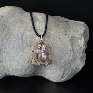 Può includere: Un ciondolo a forma di rana in conchiglia marrone e bianca con una pietra preziosa rosa sul petto, appeso a un cordoncino nero.