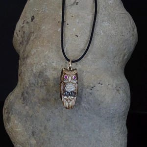 Op de afbeelding: Een ketting met hanger met een gebeeldhouwde uil-bedel met robijnkleurige ogen. De uil hangt aan een zwart koord. De hanger heeft een lichtbruine kleur met gouden accenten. De achtergrond is een lichtgrijze steen.