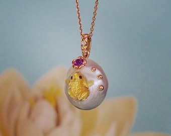 Könnte beinhalten: Eine zarte goldene Halskette mit einem Perlenanhänger, der eine kleine gelbe Frosch und einen rosafarbenen Edelstein zeigt. Der Anhänger ist von kleineren Perlen umgeben.