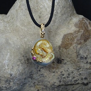 Puede incluir: Collar con colgante de oro con un diseño detallado de dragón. El colgante está suspendido de un cordón negro y tiene una fianza de oro con un pequeño diamante. Una pequeña gema rosa está engastada en el colgante.