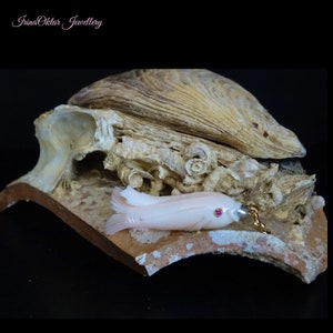 Op de afbeelding: Een delicate roze koraalvis-hanger met een robijnoog, rustend op een stuk gefossiliseerde schelp. De hanger is bevestigd aan een gouden ketting.