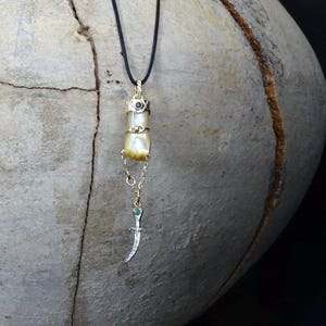Könnte beinhalten: Eine goldfarbene Anhängerkette mit einem Perlen- und Dolch-Anhänger. Der Anhänger hat eine schwarze Kordel, eine goldfarbene Oberseite mit einem schwarzen Stein und eine perlenartige Perle. Ein kleiner Dolch-Anhänger mit einem blauen Stein hängt darunter.