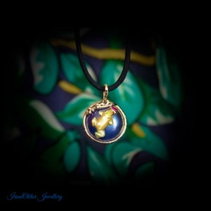 Könnte beinhalten: Eine goldene Halskette mit einem kleinen, gelben Frosch in einer blauen Glaskugel. Der Frosch hat einen roten Rubin auf dem Kopf. Die Kette ist an einem schwarzen Band.