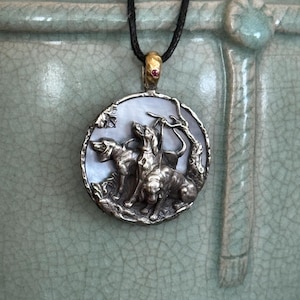 Puede incluir: Un collar con un colgante de plata redondo con una incrustación de nácar que presenta una escultura detallada de tres perros. El colgante está unido a un cordón negro.