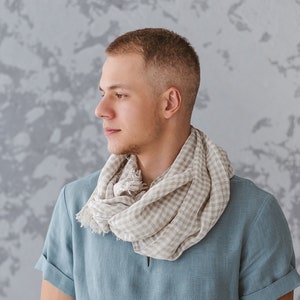 May include: A man wearing a light blue linen shirt with a beige and white checkered scarf around his neck. He is standing in front of a textured gray wall.