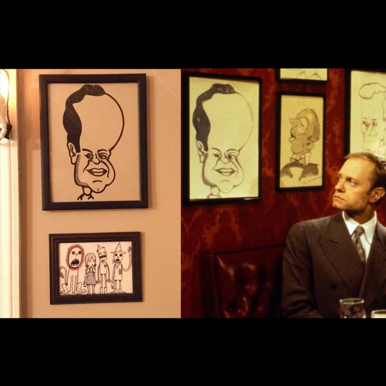 May include: A man in a suit stands in front of a wall with framed drawings. The drawings are caricatures of people with large heads. The man is looking at the drawings with a thoughtful expression.