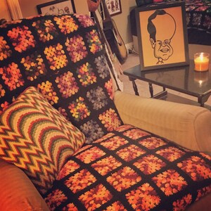 May include: A brown and beige crocheted throw blanket with a floral pattern is draped over a brown armchair. The armchair has a beige cushion with a brown and orange crocheted throw pillow on top. A small wooden table with a black top is in the background. A framed cartoon drawing of a person with a large head is on the table.