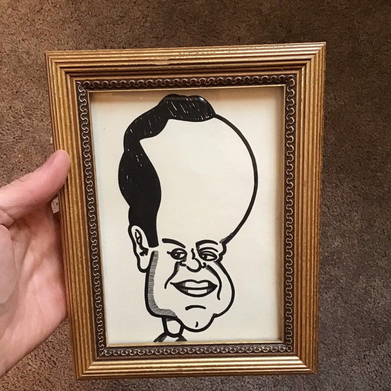 May include: A black and white caricature drawing of a person with a large head and a small body, framed in a gold frame.