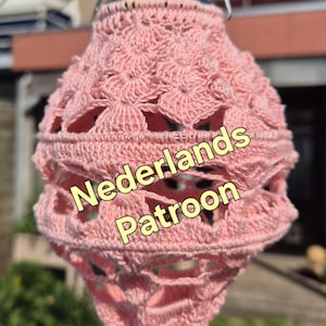 Op de afbeelding: Handgemaakte roze gehaakte lantaarn met ingewikkeld openwerk en een decoratieve bodem. Opgehangen aan een metalen haak. De tekst "Nederlands Patroon" is zichtbaar.
