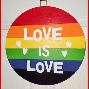 May include: A round wooden sign with a rainbow design and the words "LOVE IS LOVE" in white letters. There are also small white hearts on the sign. The sign is hanging from a red hook.