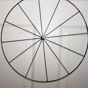 May include: A black metal wreath frame with 12 spokes. The frame is hanging from a yellow hook.
