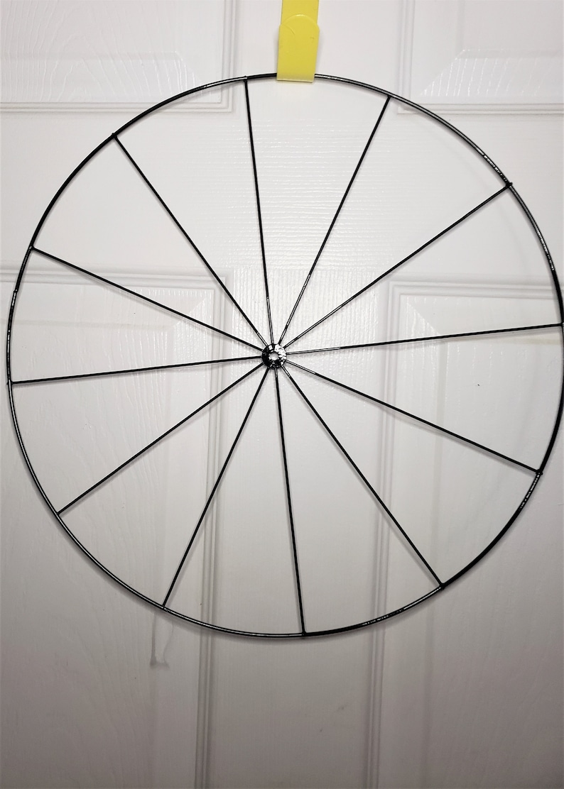May include: A black metal wreath frame with 12 spokes. The frame is hanging from a yellow hook.