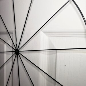 May include: A black metal circular wreath with spokes radiating from the center. The wreath is against a white wall.