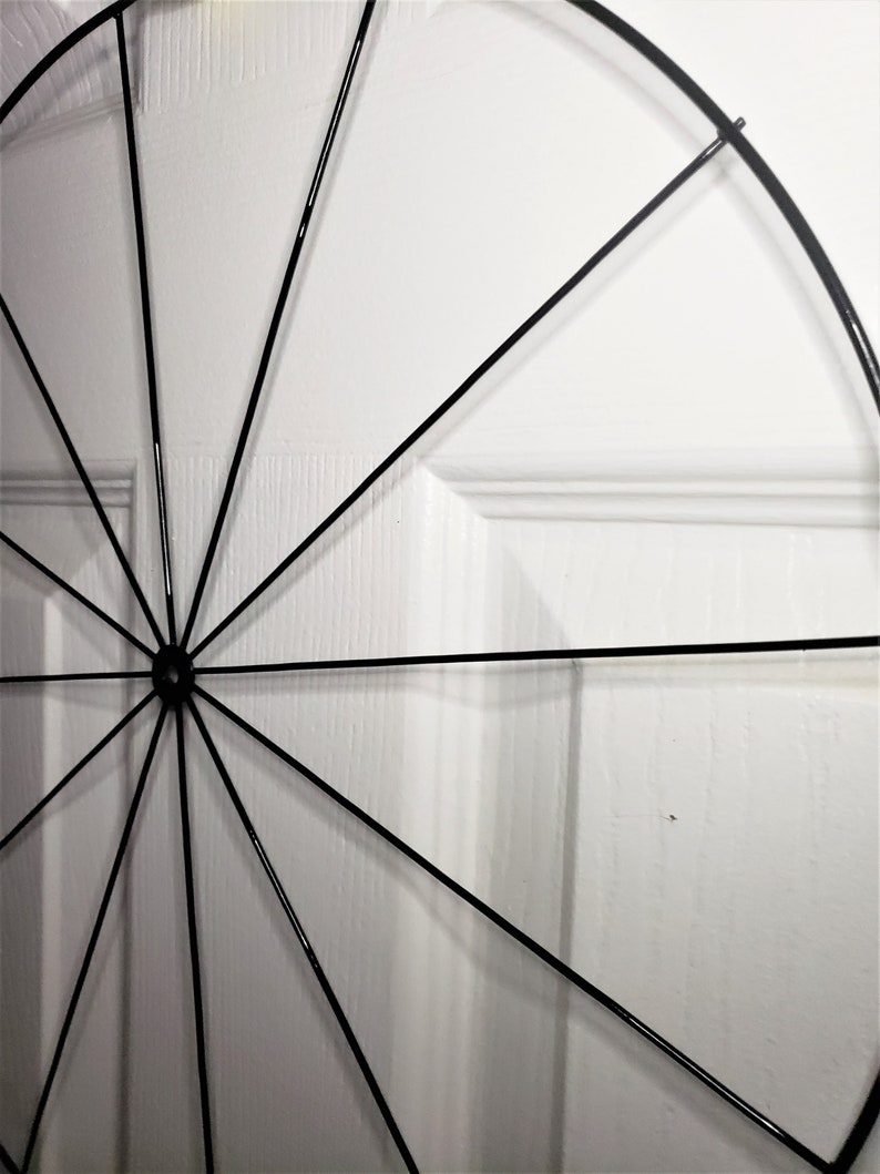 May include: A black metal circular wreath with spokes radiating from the center. The wreath is against a white wall.
