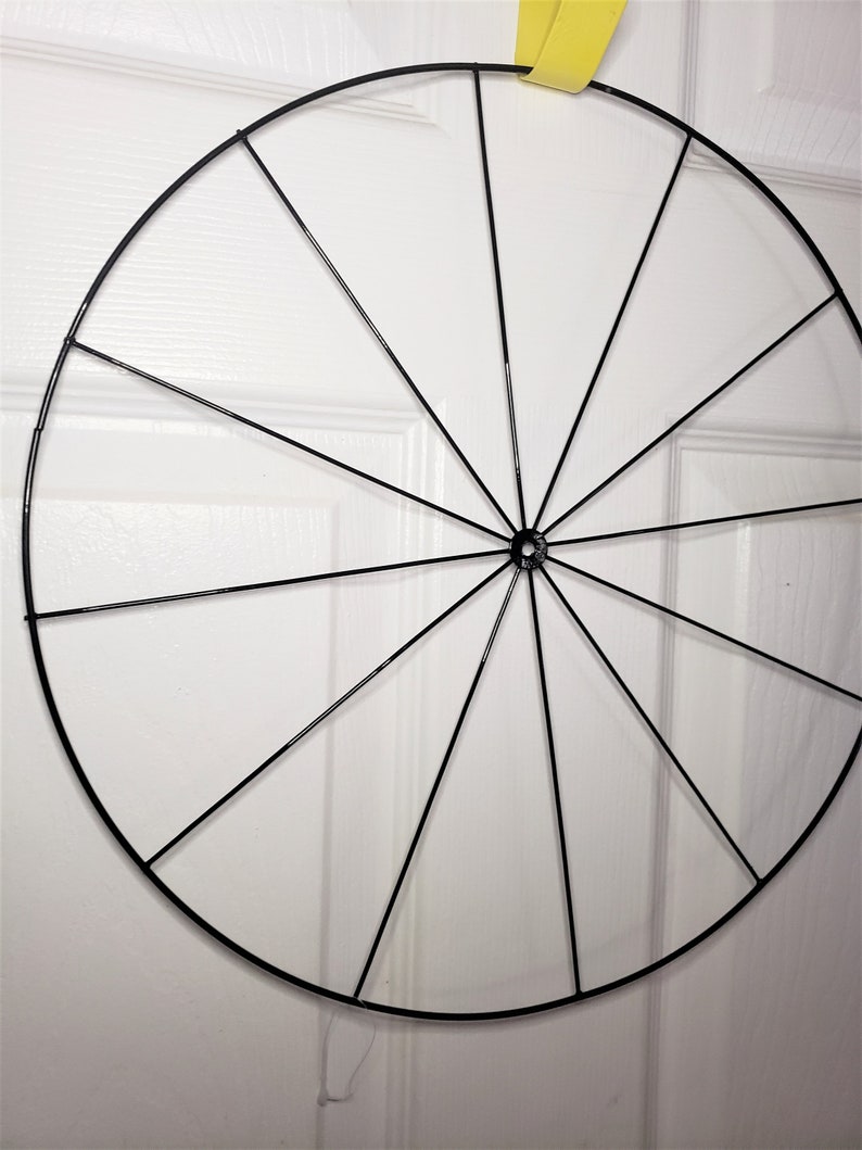May include: A black metal wreath frame with 12 spokes. The frame is circular and is hanging from a yellow ribbon.