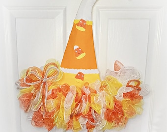 Könnte beinhalten: Ein Halloween-Kranz mit einem Hexenhut aus orangefarbenem und weißem Stoff mit Mais-Karamell-Designs. Der Hut ist von einem flauschigen orangefarbenen, gelben und weißen Netz umgeben.