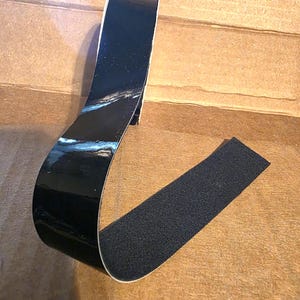 May include: A roll of black, glossy tape with a contrasting strip of black hook-and-loop fastener material. The tape is curved, showing the adhesive side and the textured surface of the hook-and-loop material. The background is a light brown surface.