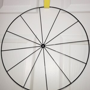 May include: A black metal wreath frame with 12 spokes. The frame is circular and is hanging from a hook.