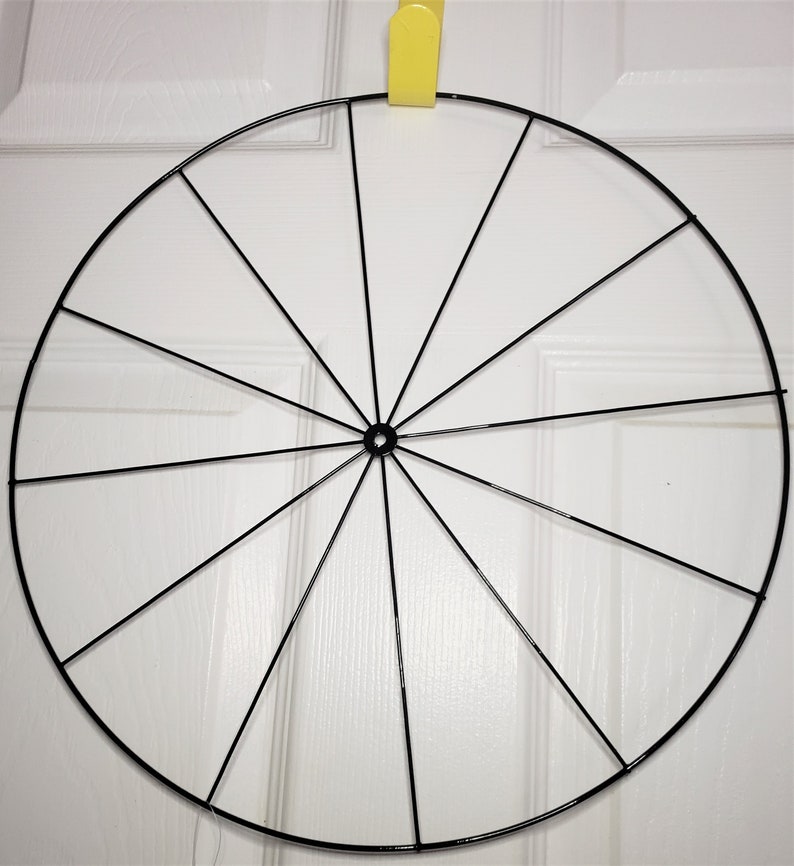 May include: A black metal wreath frame with 12 spokes. The frame is circular and is hanging from a hook.