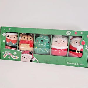 May include: A box of five plush toys with a Christmas theme. The toys are a Christmas tree, Santa Claus, a reindeer, a grandma, and an elf. The box is green with the words "Original Squishmallows" and "Seasonal Squad" printed on it.