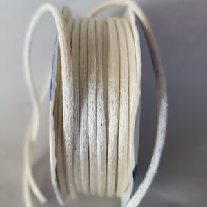 May include: A spool of off-white, sparkly cord. The cord is tightly wound around a cardboard core, with a few loops extending outwards. The cord has a subtle sheen, suggesting a satin or similar finish. Ideal for crafting and jewelry making.