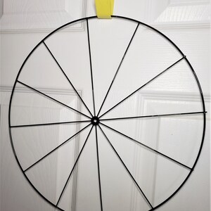 May include: A black metal wreath frame with 12 spokes. The frame is circular and is hanging from a yellow hook.