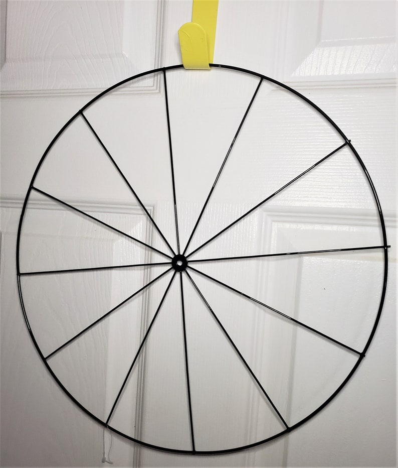 May include: A black metal wreath frame with 12 spokes. The frame is circular and is hanging from a yellow hook.