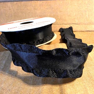 May include: A spool of black ribbon with a ruffled edge. The ribbon is made of a woven material and is partially unraveled, showing the texture and detail. The spool has text that reads "TRIM - ORNER - ADORNO".