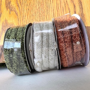 May include: Three spools of decorative ribbon in clear plastic packaging. The colors are green, white, and copper. Each spool has a textured, looped design. A small pearl is attached to the front of the copper and white spools.