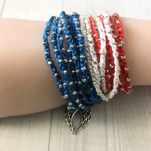 May include: A stack of colorful bracelets with silver beads, featuring red, white, and blue colors. The bracelets are tied together with a silver charm in the shape of angel wings.