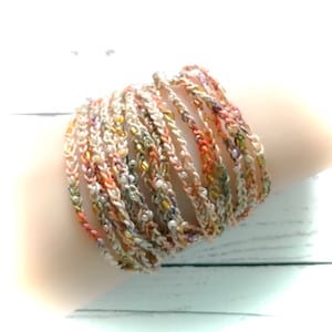 May include: A multi-strand bracelet made of crocheted cord and beads in shades of beige, peach, and gold. The bracelet is layered and has a bohemian style.