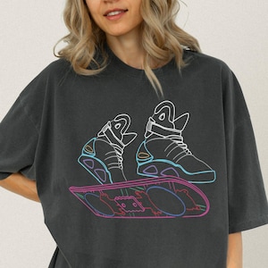 May include: Dark gray t-shirt featuring a neon outline design of futuristic sneakers and a hoverboard. The graphic is in shades of blue, pink, and yellow. The shirt has a relaxed fit and short sleeves.