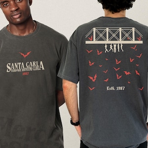 May include: Dark gray t-shirt with the text "SANTA CARLA VAMPIRE HUNTING CLUB 1987" on the front and a bridge design with bats on the back. The back of the shirt also has the text "Est. 1987".