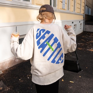 Puede incluir: Sudadera color crema con la palabra "SEATTLE" impresa en letras grandes, azules y estilizadas en la espalda, con una línea verde en el medio. La sudadera se usa con una falda negra y una gorra de béisbol marrón.