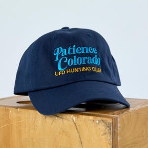 Pode incluir: Boné de beisebol azul marinho com as palavras "Patience Colorado" bordadas em azul claro e "UFO HUNTING CLUB" em amarelo. O boné está sobre uma superfície de madeira.
