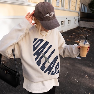 May include: A beige sweatshirt with the word "DALLAS" in navy blue, a brown cap with "La Paris" text, and a black purse. The sweatshirt has a white undershirt. The cap is brown. The person is holding a coffee drink.