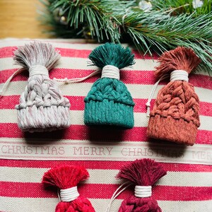 May include: Five handmade knitted Christmas ornaments shaped like winter hats in various colors, including red, green, white, brown, and burgundy. The ornaments are displayed on a red and white striped background.