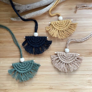 May include: Four macrame wall hangings with fringe, each in a different color: mustard yellow, black, green, and beige. Each hanging has a white wooden bead at the top.