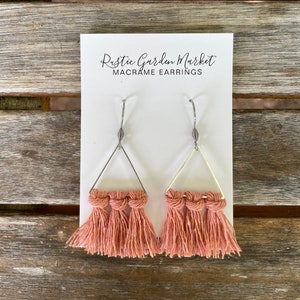 May include: A pair of pink macrame earrings with triangle-shaped silver hoops. The earrings are labeled "Rustic Garden Market Macrame Earrings".