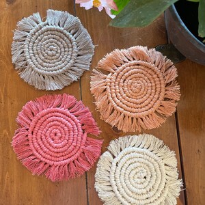May include: Four handmade crocheted coasters with fringe. The coasters are in shades of beige, peach, pink, and white.