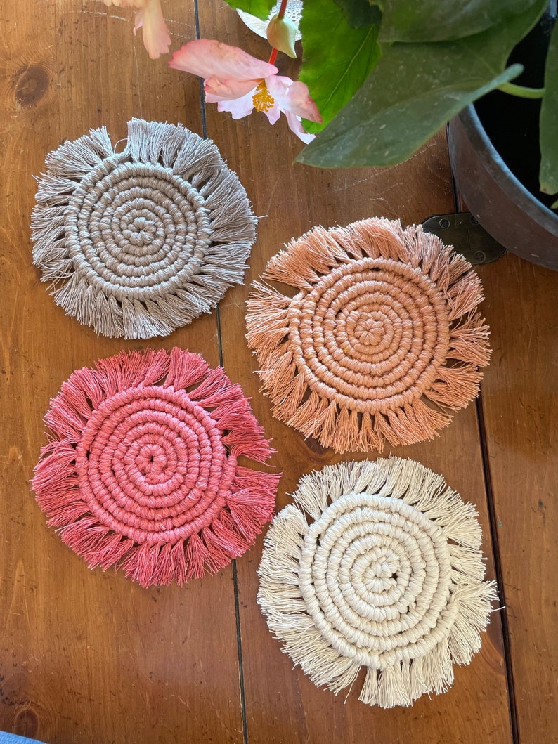 May include: Four handmade crocheted coasters with fringe. The coasters are in shades of beige, peach, pink, and white.