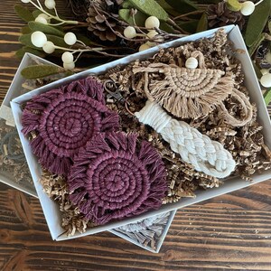 May include: A gift box filled with handmade macrame items. The box contains two burgundy coasters, a white macrame keychain, and a beige macrame wall hanging.