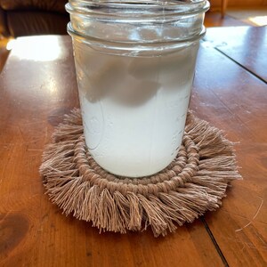 May include: A glass jar filled with a white liquid, sitting on a brown crocheted coaster with fringe.