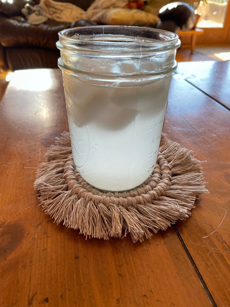 May include: A glass jar filled with a white liquid, sitting on a brown crocheted coaster with fringe.